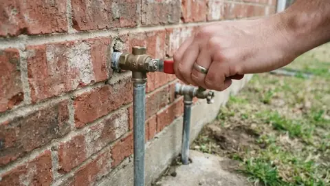 Hand turning a shutoff valve on a water line against a brick wall